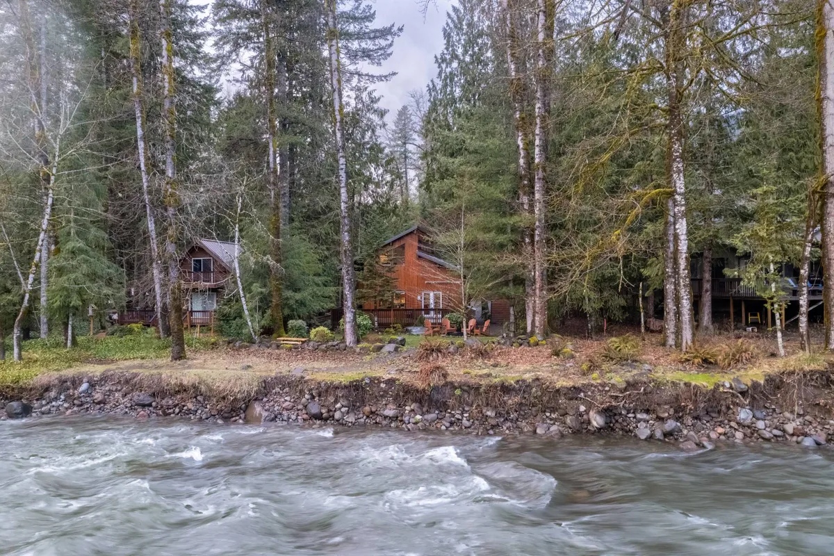This image provides a wide view of the log cabin nestled by a river, framed by tall trees and featuring a patio seating area. It conveys a sense of retreat and connection to nature, while the river suggests opportunities for peaceful contemplation or group activities by the water.