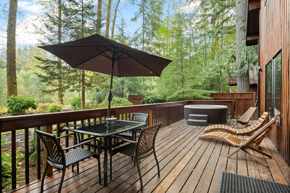 A spacious outdoor setting featuring a dining table, hot tub, and lounge chairs set against a backdrop of tall trees. The area is ideal for relaxing after a day of outdoor activities, socializing, or dining al fresco while taking in sunset views over the forest.