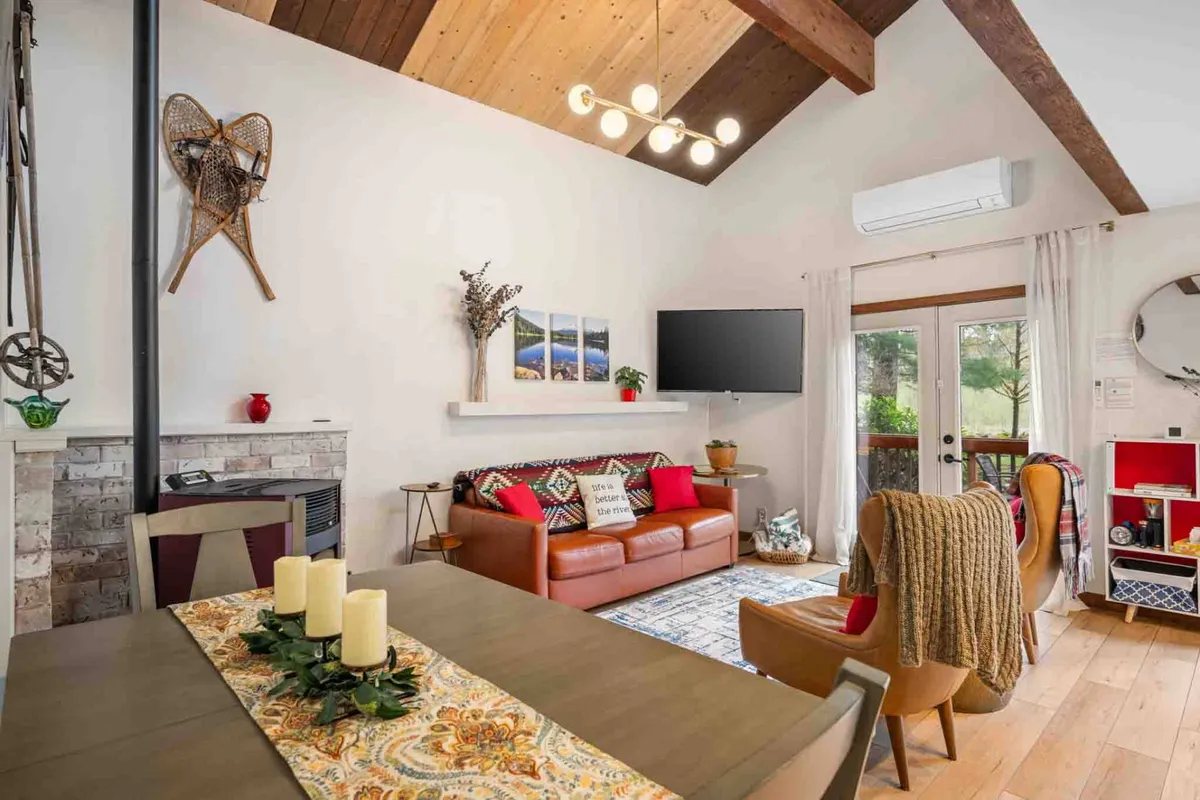 A spacious, open-concept living and dining area featuring rustic wooden beams, a comfortable leather sofa with colorful throw pillows, and a large wooden dining table set with candles. Expansive windows illuminate the space with natural light and lead out to an inviting outdoor area.