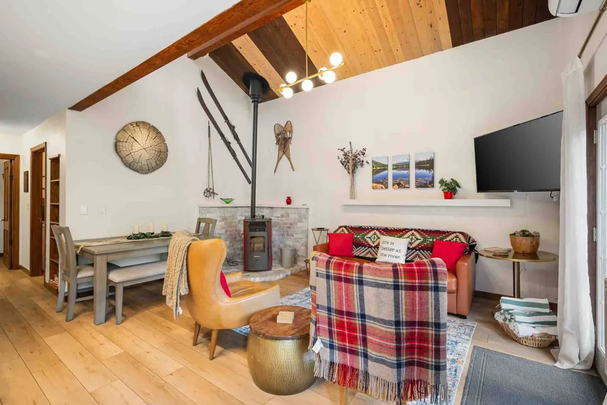 A warm, welcoming interior that features a colorful couch near a pellet fireplace, a dining table for shared meals, exposed wooden beams, and tasteful wall decor. The area feels both practical and homely, perfect for cozy winter evenings or socializing with family and pets.