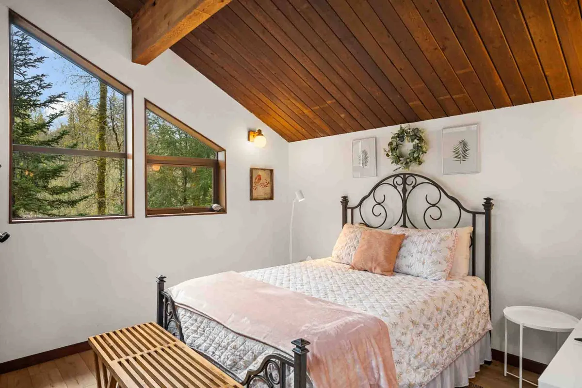 A tranquil bedroom featuring a bed with floral bedding, unique windows allowing natural light to stream in, decorative wall art, and a wooden ceiling. The scene evokes a sense of peacefulness and invites relaxation, making it ideal for restful nights.