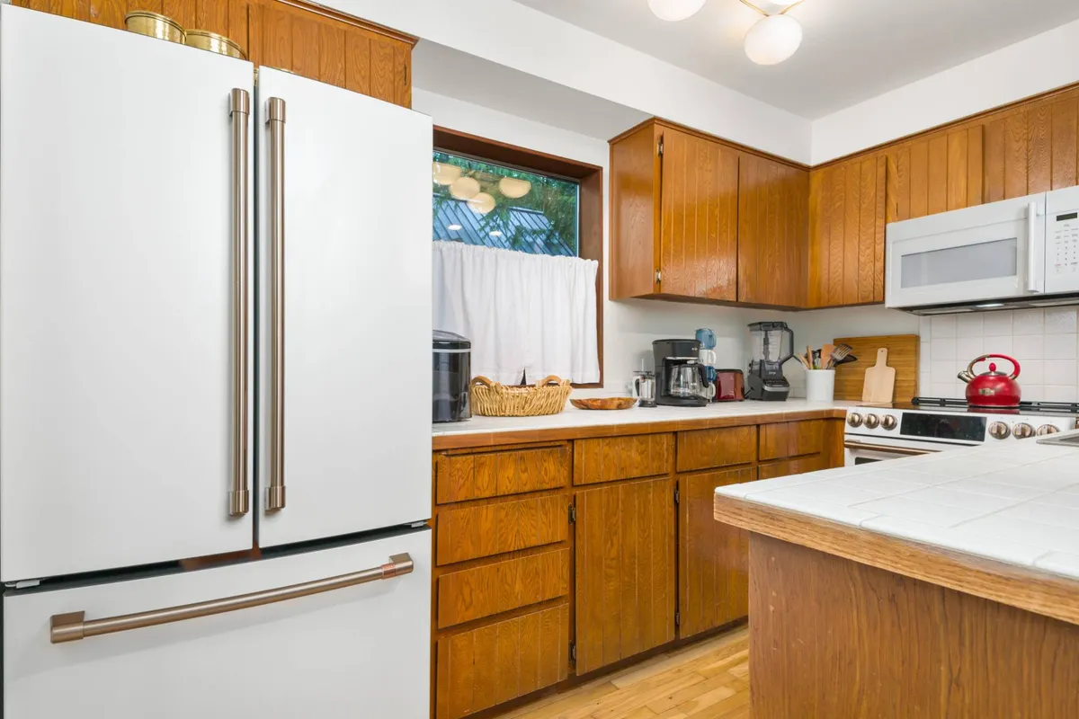 A homey kitchen with white refrigerator, wooden cabinets, countertop appliances, and a window with curtains. The kitchen is ready for family gatherings, baking, and preparing meals, emphasizing the practicality and completeness of amenities.
