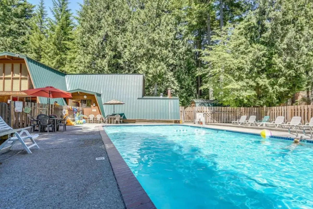 A sunlit swimming pool area with lounge chairs and umbrellas, surrounded by trees and a community building. The setting is perfect for summer relaxation, swimming, and enjoying the seasonal amenities offered to guests.