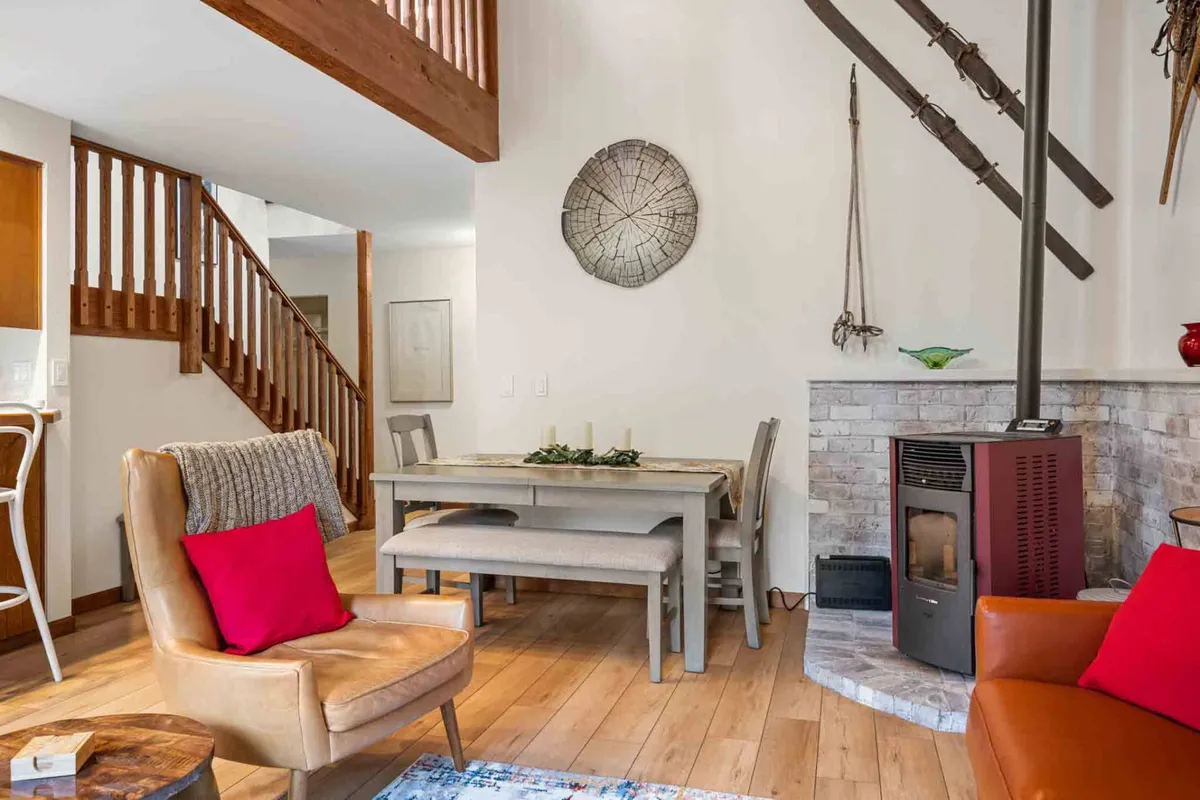 A cozy interior view showing a large wooden dining table set for a meal, a warm fireplace, a wooden staircase, and a colorful armchair. The space exudes warmth and is ideal for family gatherings and laughter-filled meals.