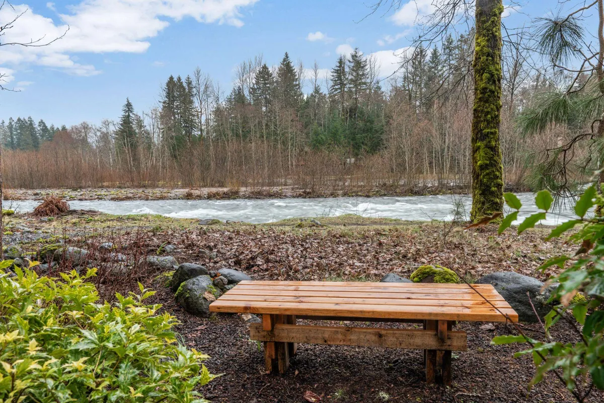 A serene riverside scene featuring a wooden bench overlooking the flowing river, surrounded by trees and a rock border. The setting is perfect for picnicking, nature observation, or simply relaxing by the water’s edge.