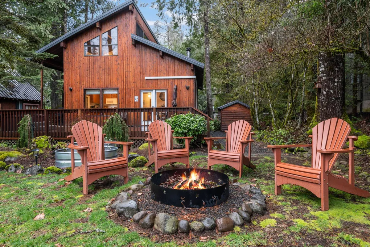 The image showcases a cozy wooden cabin nestled among towering trees on the banks of the Sandy River. Adirondack chairs circle a glowing fire pit, inviting guests to gather for warm evenings under the stars, surrounded by the tranquil beauty of the Pacific Northwest wilderness.