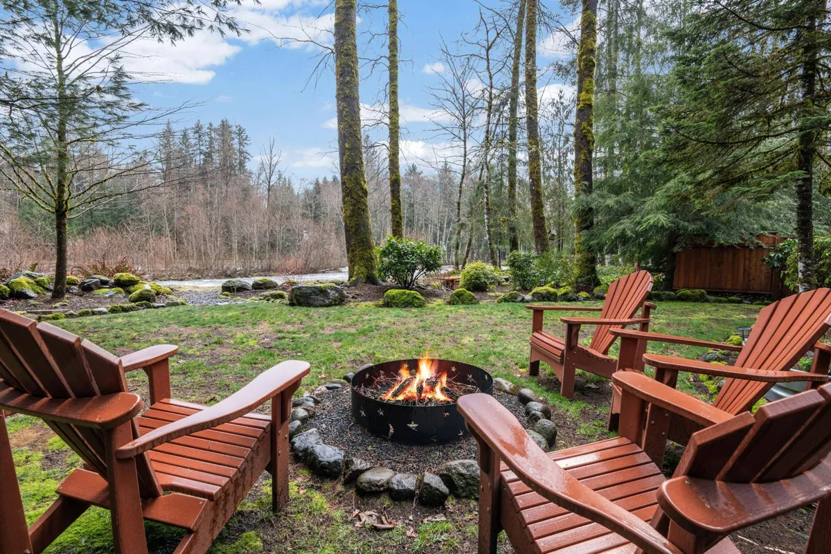 A cozy fire pit surrounded by Adirondack chairs overlooks the river and is framed by trees. The scene captures the essence of evening gatherings, marshmallow toasting, and stargazing by the water.