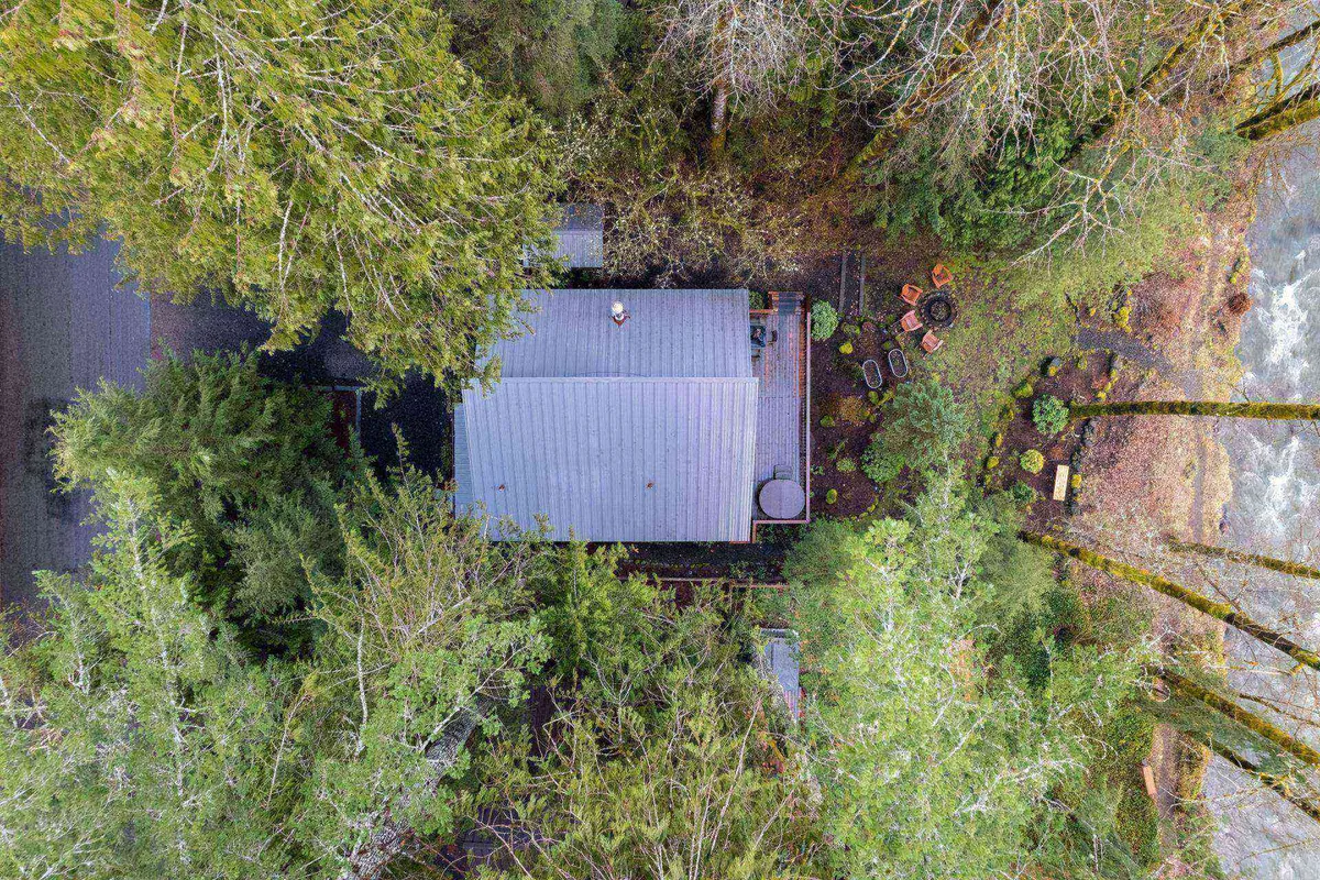 This image shows a house nestled among trees with a clear view of the river and an inviting outdoor seating area. The natural setting emphasizes the close connection to the river and the generous outdoor space for relaxation or play.